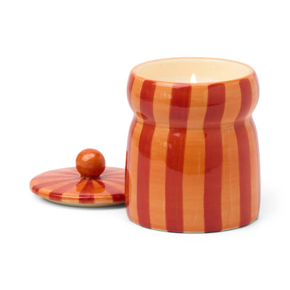 A Spiced Apple - Cabana 6.5 oz Candle burns inside a striped orange and red ceramic jar, with its matching lid placed beside it.