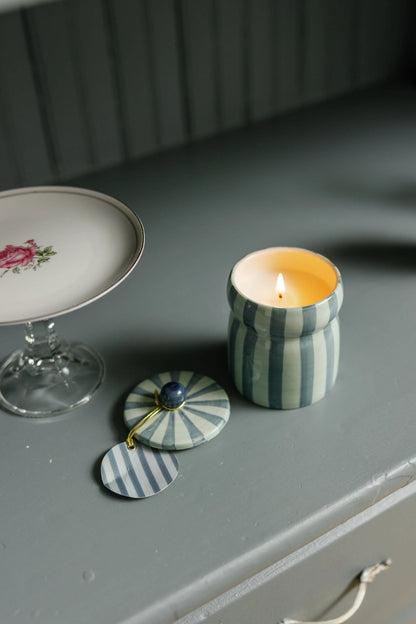 A lit Cabana 6.5 oz Warm Cider candle rests on a gray surface with its ceramic lid placed beside it, next to a glass dish featuring a floral design. The hand-painted cabana vessel enhances the scene with charm, while the mulled cider fragrance fills the air with inviting warmth.