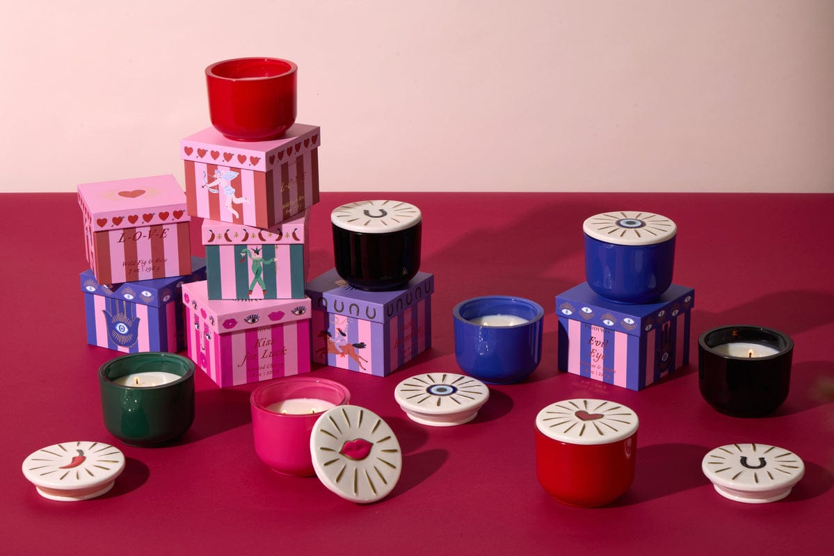 Colorful candles in red, green, blue, and black containers with illustrated lids and matching decorative boxes are arranged on a red and pink surface against a pale pink background.
