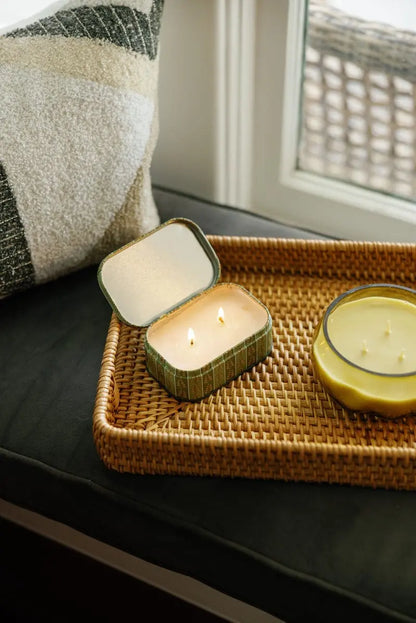 A wicker tray holds an open Smoked Pistachio - Chérie 4.5 oz Tin Candle with two wicks alongside a round yellow candle. The tray rests on a dark cushion near a window, with a patterned pillow in the background, infusing the space with an inviting gourmand fragrance.