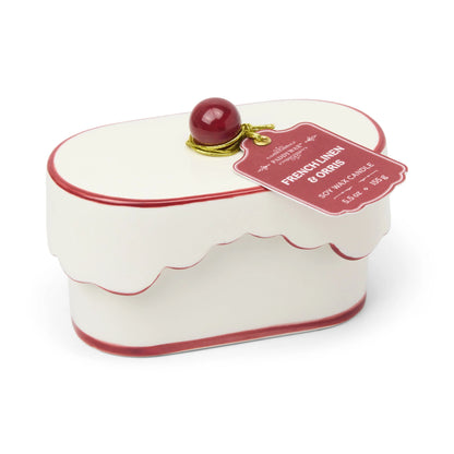 White and red scalloped tin box with a round red knob on top, resembling a cake. A pink tag attached reads French Linen & Orris - Chérie 5.5 oz Ceramic Candle, reminiscent of the elegance found in a porcelain vessel or the soothing fragrance of French linen blends.