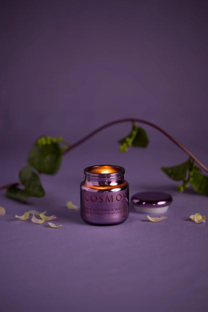 A lit Wisteria & Willow - Chroma 2.5 oz Candle in an electroplated glass metallic jar sits on a purple surface with flower petals and a leafy vine in the background. Part of the Chroma Collection candles, its lid rests beside the candle.
