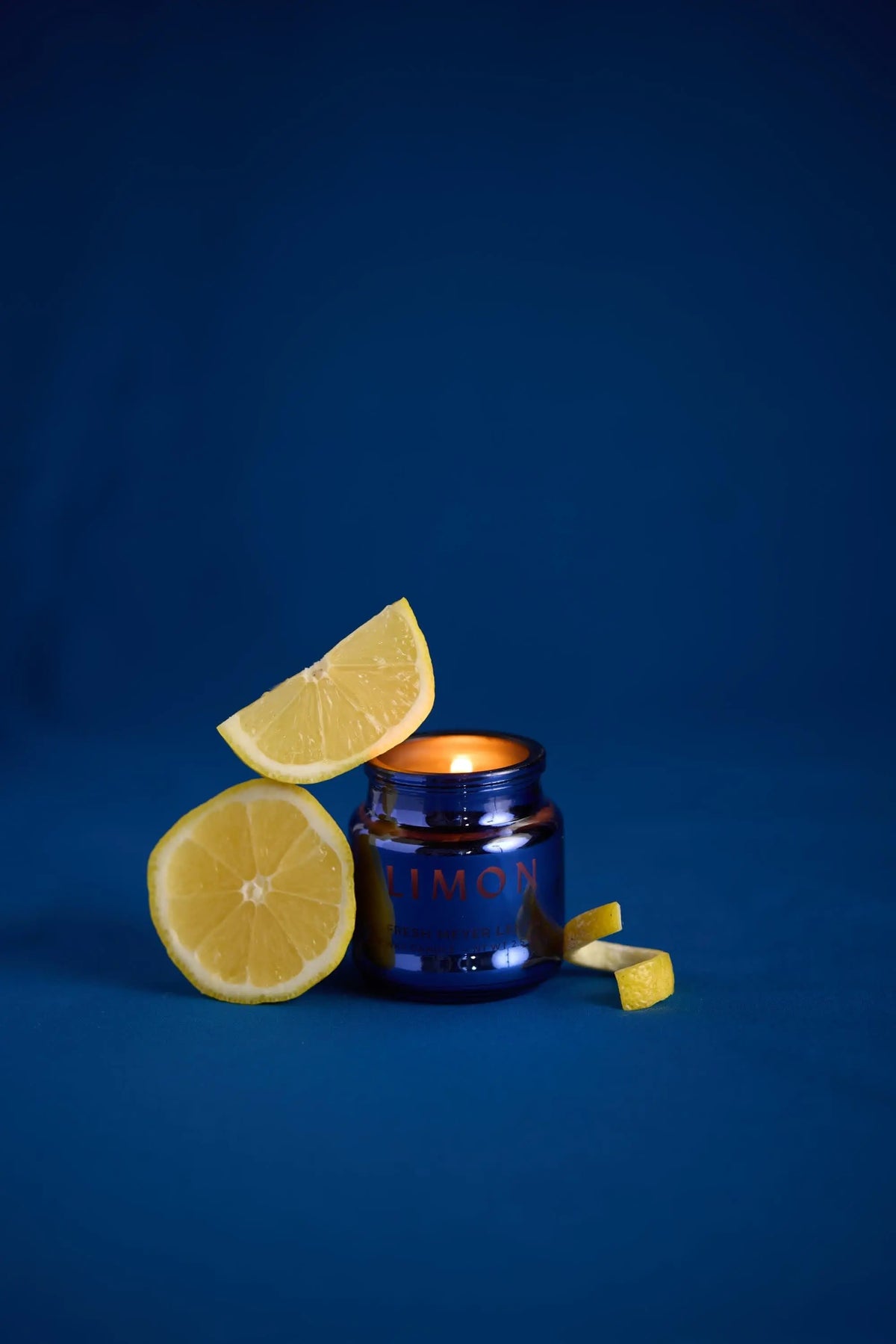 The Meyer Lemon - Chroma 2.5 oz Candle glows in its dark blue electroplated glass jar, placed on a blue surface surrounded by a lemon half, a wedge balanced on the jar, and two small pieces of lemon arranged nearby.