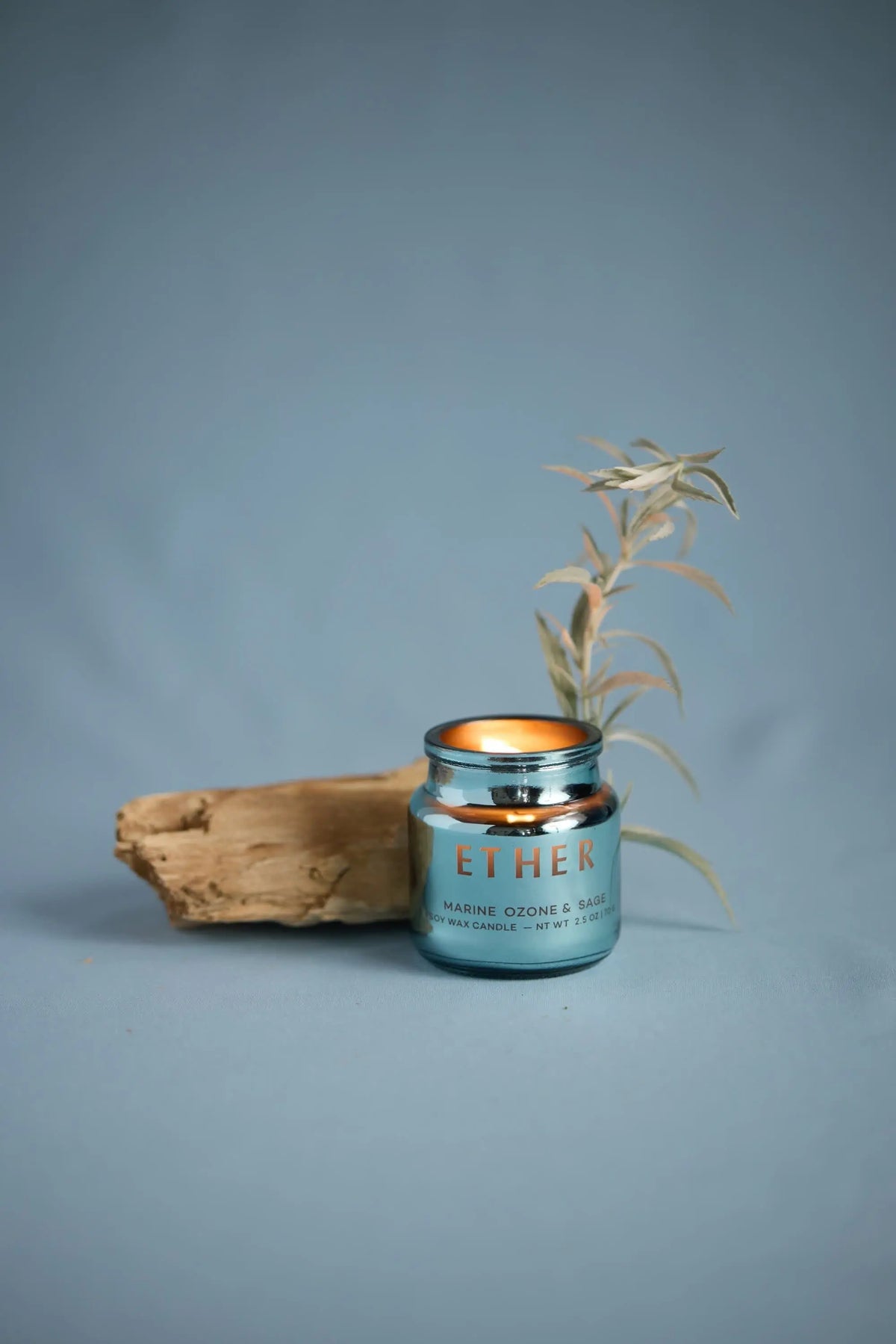 A lit Sea Salt & Sage - Chroma 2.5 oz Candle in a blue jar sits beside a piece of driftwood and a pale leafy branch on a light blue background.