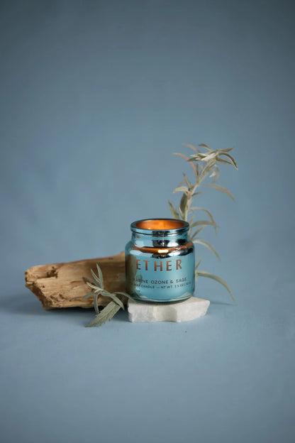 A blue Sea Salt & Sage - Chroma 2.5 oz candle jar sits on a white stone with a beige wood piece and a light green leafy twig beside it, set against a plain blue background. Part of the Chroma collection, this candle is housed in electroplated glass for an elegant display.