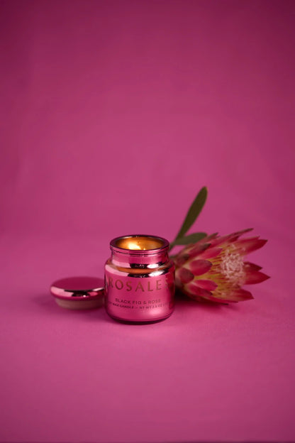 The Saffron Rose - Chroma 2.5 oz Candle, with a lit flame, sits next to its lid and a protea flower on a pink background.