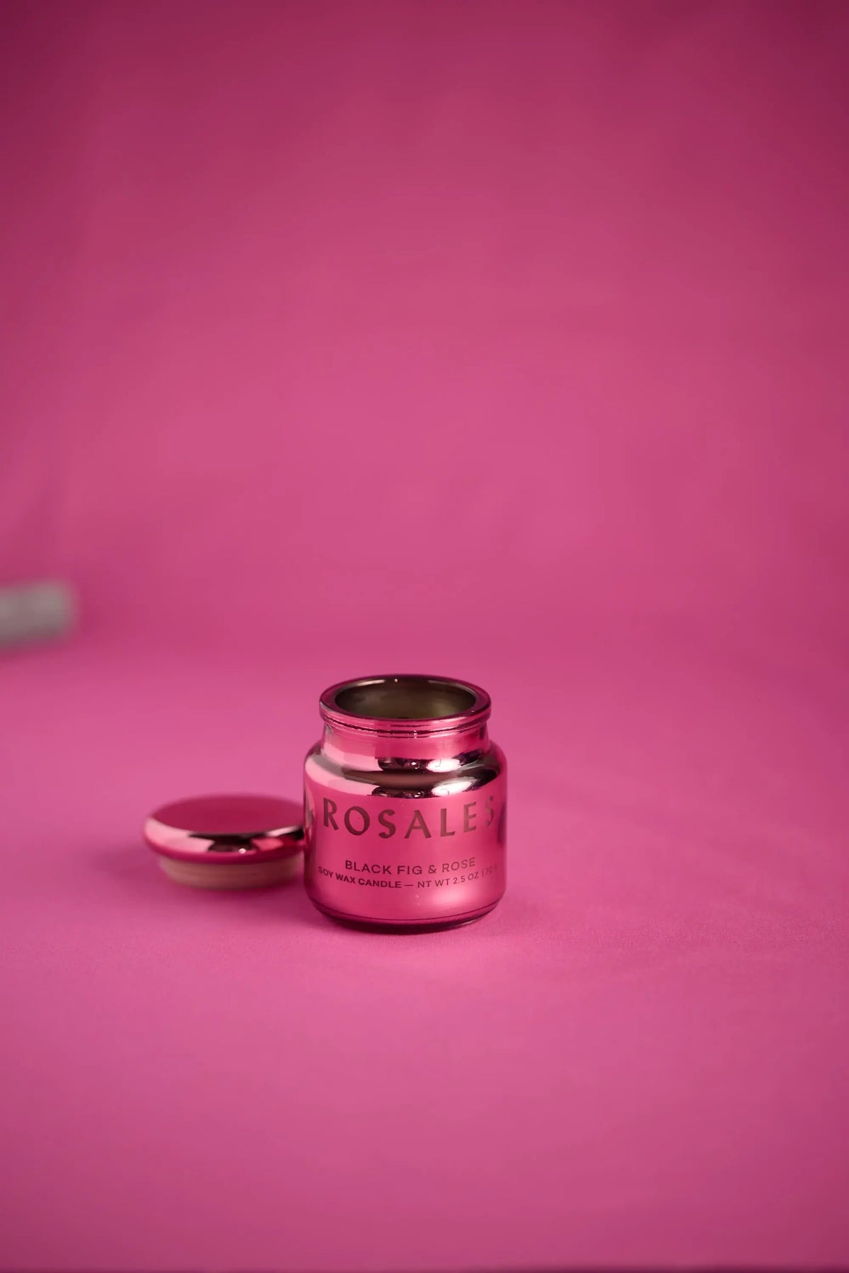 A metallic pink jar labeled Saffron Rose, from the Chroma 2.5 oz Candle collection, sits with its lid off on a pink surface against a matching pink background. This electroplated glass candle adds a touch of glamour while infusing the space with its luxurious Latin-inspired scent.