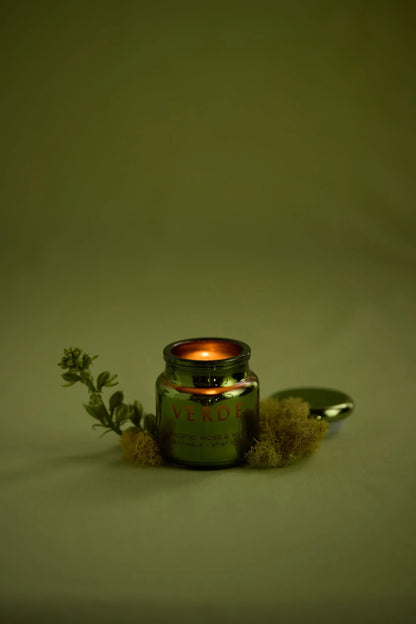 A small Pacific Moss & Mist - Chroma 2.5 oz Candle glows in an electroplated glass jar labeled VERDE, nestled among moss and greenery, with its lid set aside on a soft beige background. This Chroma candle exudes a Latin-inspired fragrance, capturing the lush vibrancy of nature.