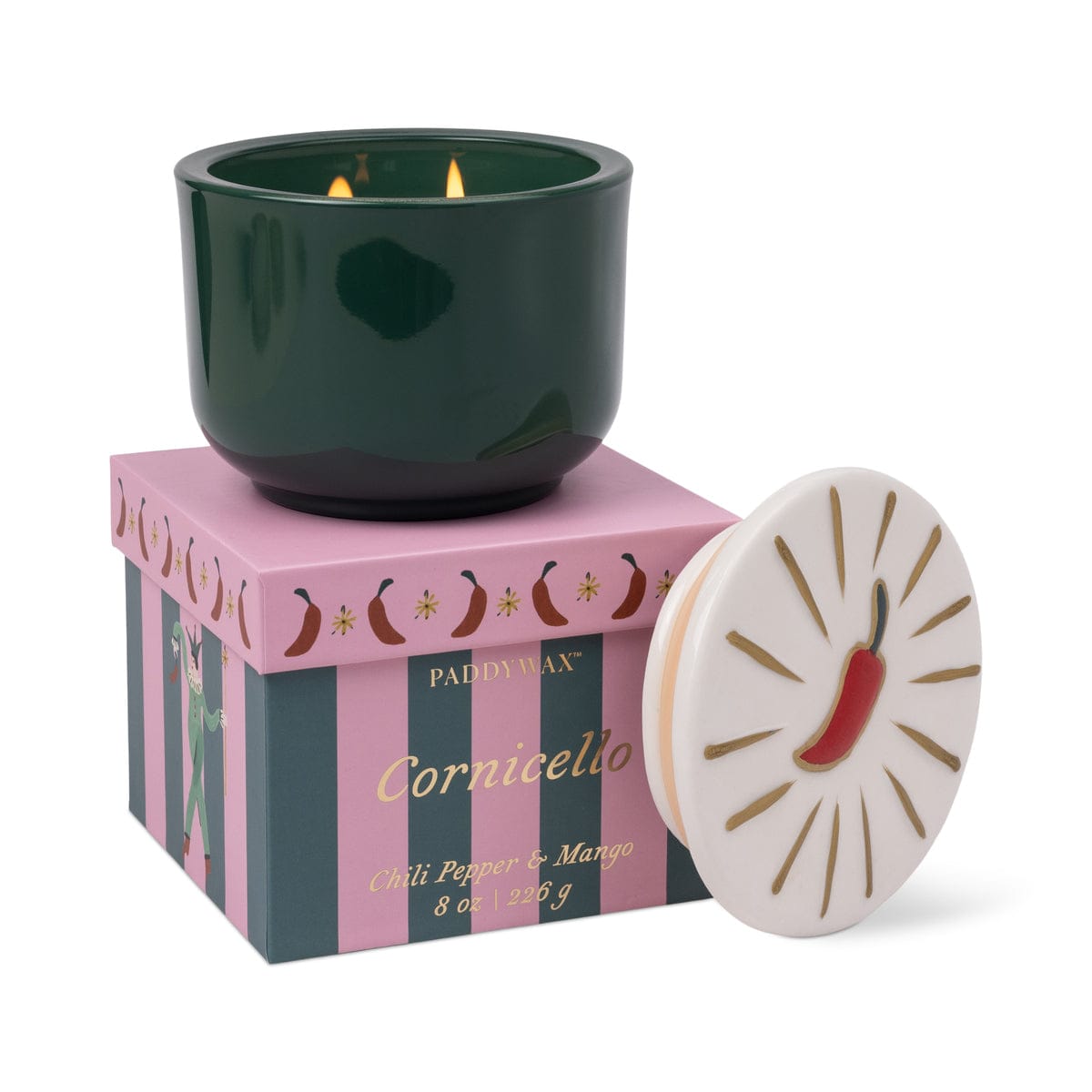 A green Chili Pepper & Mango - Charmed 8oz Candle sits in a glass cup on top of a purple and pink striped box labeled Cornicello. The white ceramic lid, featuring a red chili pepper design, rests beside it. This candle offers a delightful Chili Pepper & Mango scent.