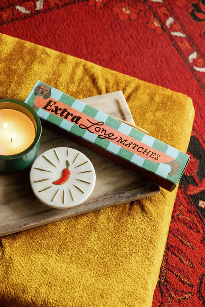 A box of Dachshund - Extra Long Boxed Matches, a lit candle, and a chili-illustrated coaster sit on a beige tray over gold fabric—perfect as a unique dachshund gift with stylish safety matches.