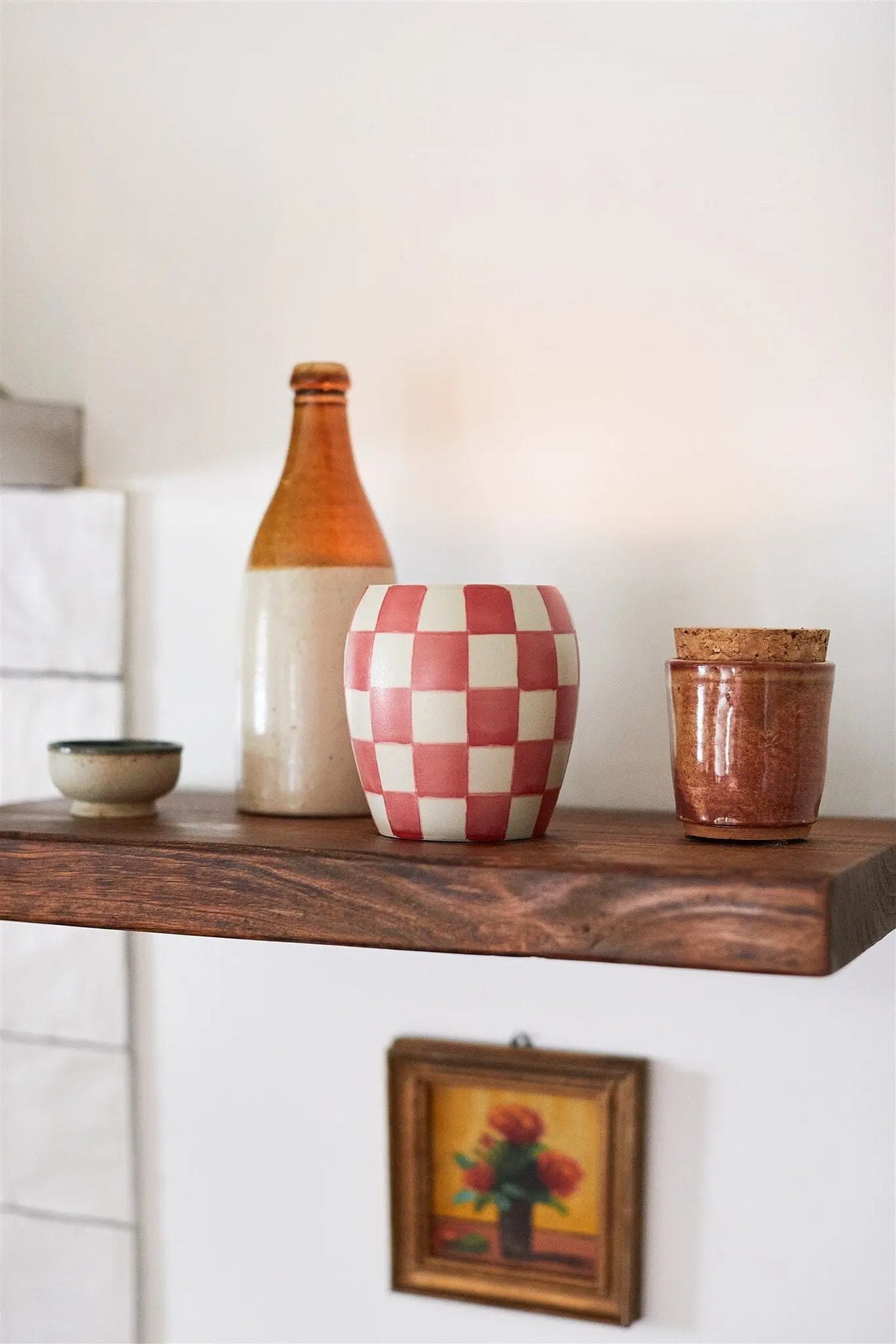 A wooden shelf displays the Rose + Santal - Checkmate Candle 11 oz, a small bowl, a two-tone bottle, hand-painted vessels, and a corked jar; below hangs a small framed floral painting.