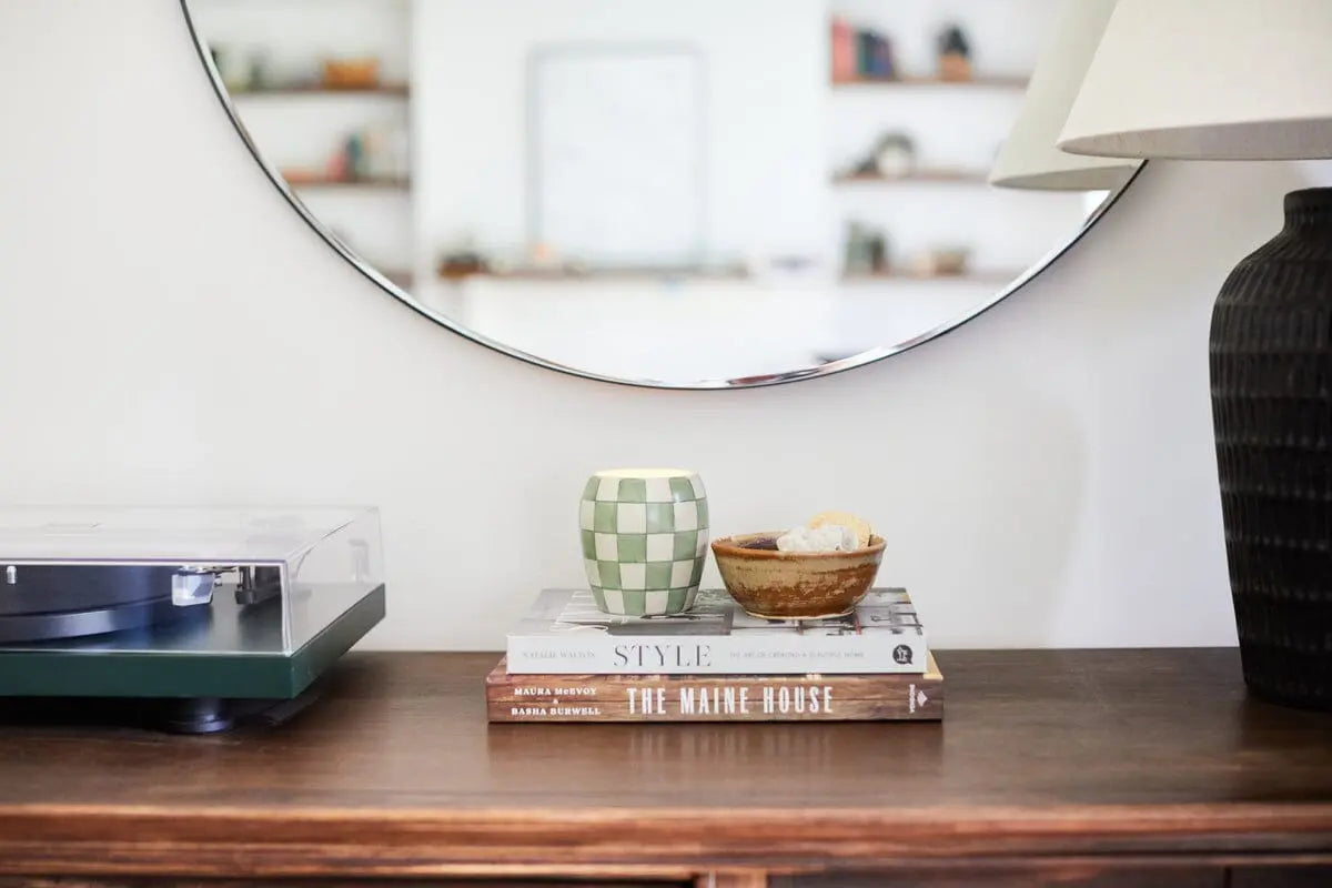 A round mirror above a wooden console displaying a record player, two stacked books, a Checkmate 11 oz Candle - Cactus Flower, a small bowl with snacks, and a black table lamp.