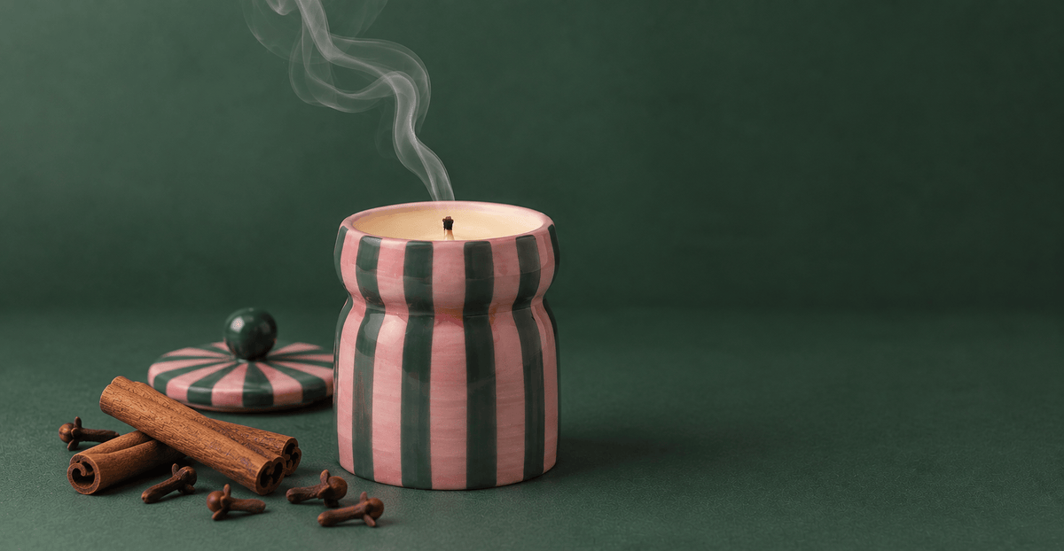 A striped jar candle with pink and dark green vertical stripes, shown lit with rising smoke on a dark green background, with cinnamon sticks and cloves beside the lid.