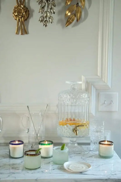 A marble countertop displays the Vanilla Rosa - La Playa 9 oz Candle, a glass dispenser with citrus-infused water, empty glasses, and decorative gold wall accents.