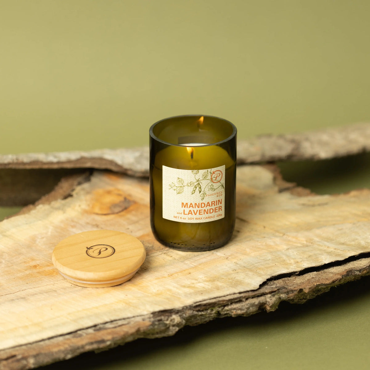 The Mandarin + Lavender - Eco 8 oz Candle, in a dark green glass jar with a round wooden lid, sits on wood against a green background—an inviting accent for sustainable living.