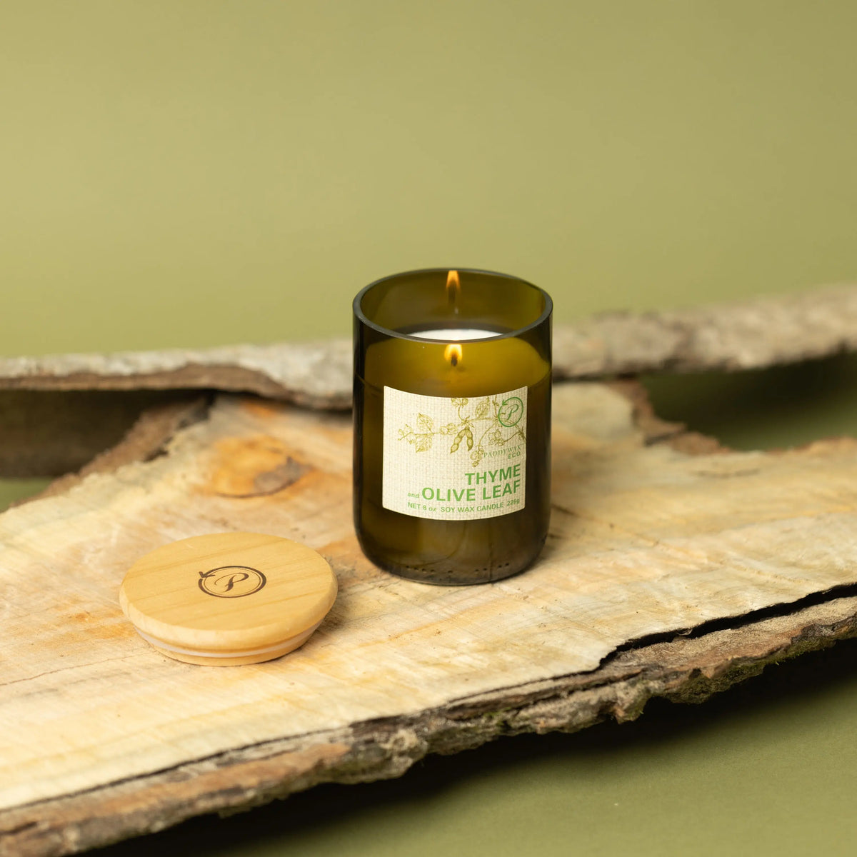 The Thyme + Olive Leaf - Eco 8 oz Candle in green glass rests on rustic wood with its wooden lid nearby, set against an olive-green background—ideal for bringing a soothing herbal scent to your space.