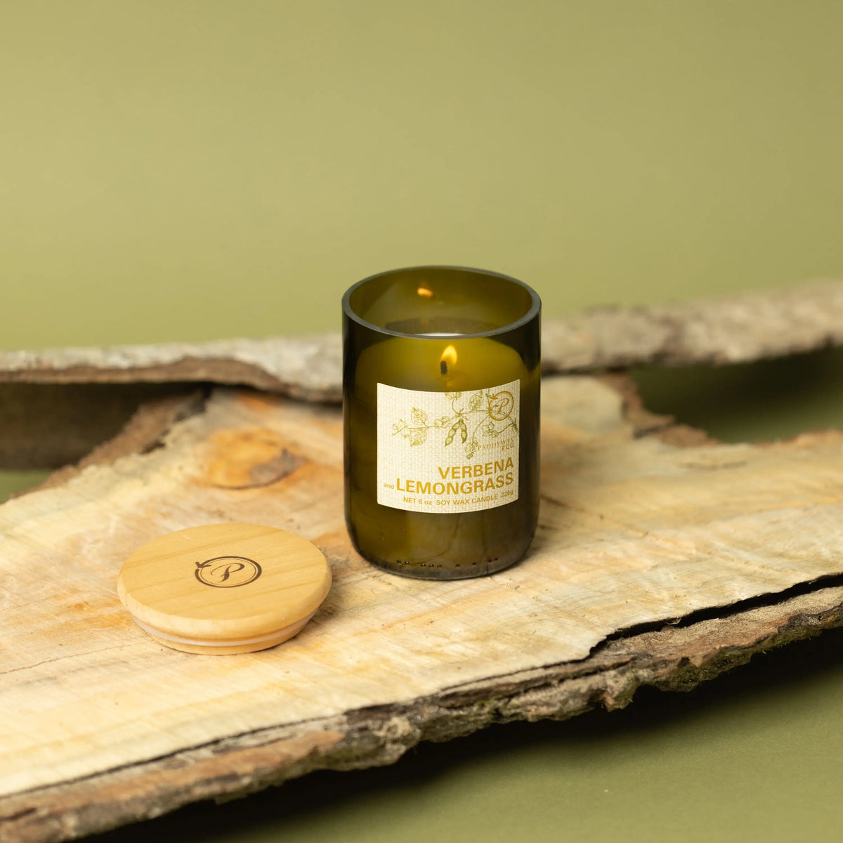 The Verbena + Lemongrass - Eco 8 oz Candle glows in a green glass jar on wood, with its wooden lid beside it. Set against a light green background with bark accents, it emphasizes the natural vibe of this eco-friendly candle.