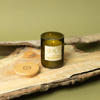 The Verbena + Lemongrass - Eco 8 oz Candle glows in a green glass jar on wood, with its wooden lid beside it. Set against a light green background with bark accents, it emphasizes the natural vibe of this eco-friendly candle.