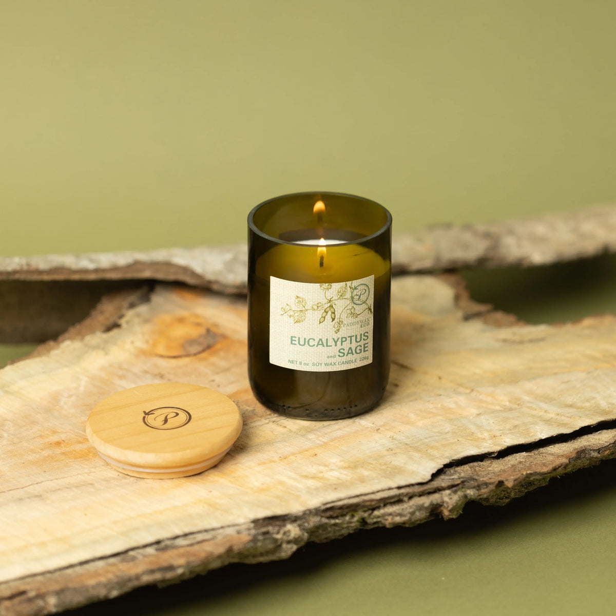 The Eucalyptus + Sage - Eco 8 oz Candle in a green recycled glass jar sits on wood with its wooden lid nearby. Green tones and tree bark pieces create a natural backdrop for this eco-friendly candle.