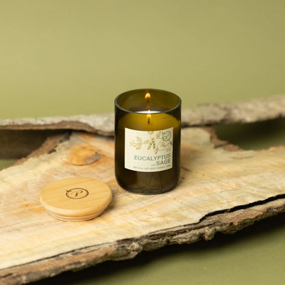 The Eucalyptus + Sage - Eco 8 oz Candle in a green recycled glass jar sits on wood with its wooden lid nearby. Green tones and tree bark pieces create a natural backdrop for this eco-friendly candle.