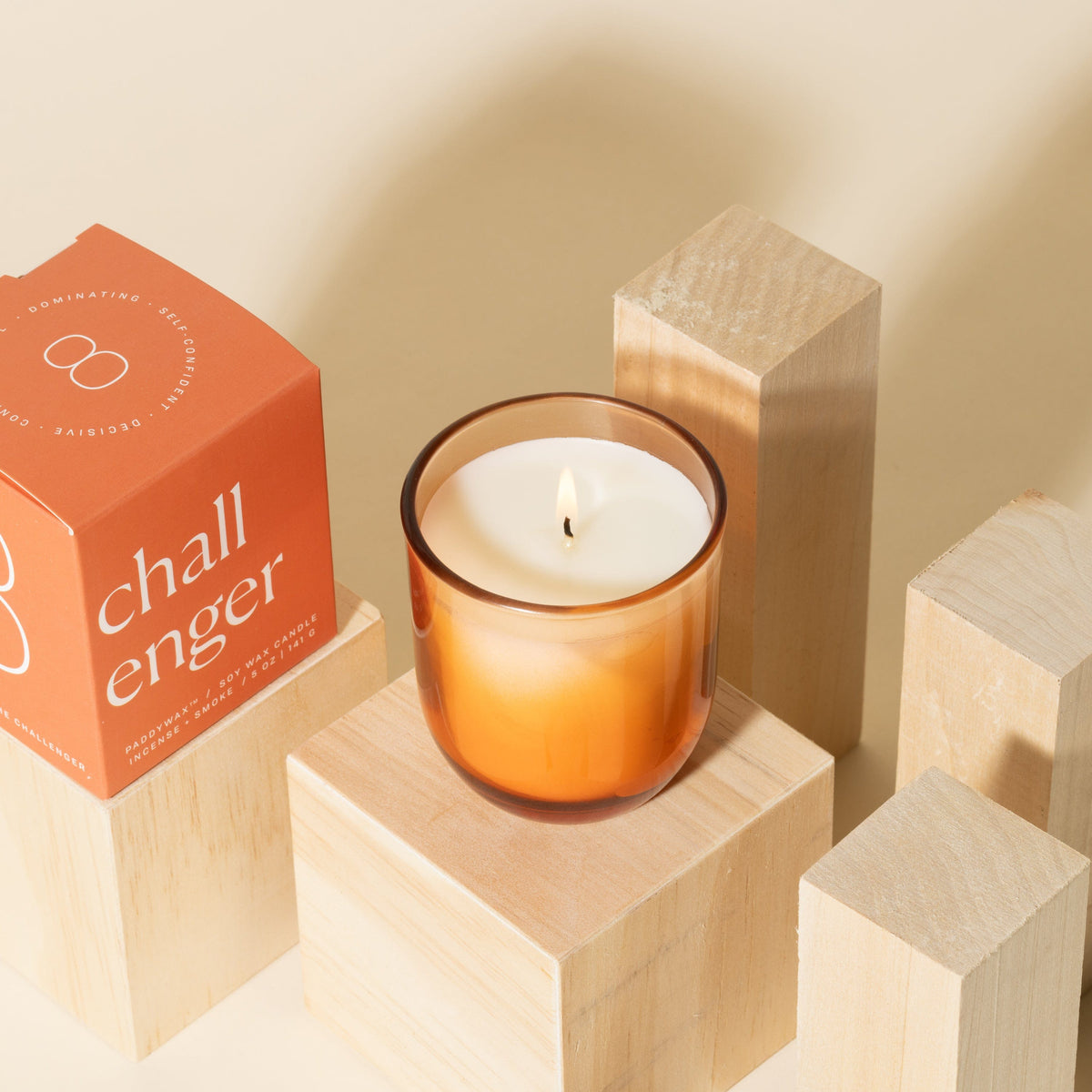 An orange glass candle sits lit on a wooden block, next to its orange box labeled challenger, surrounded by additional light wooden blocks against a beige background.