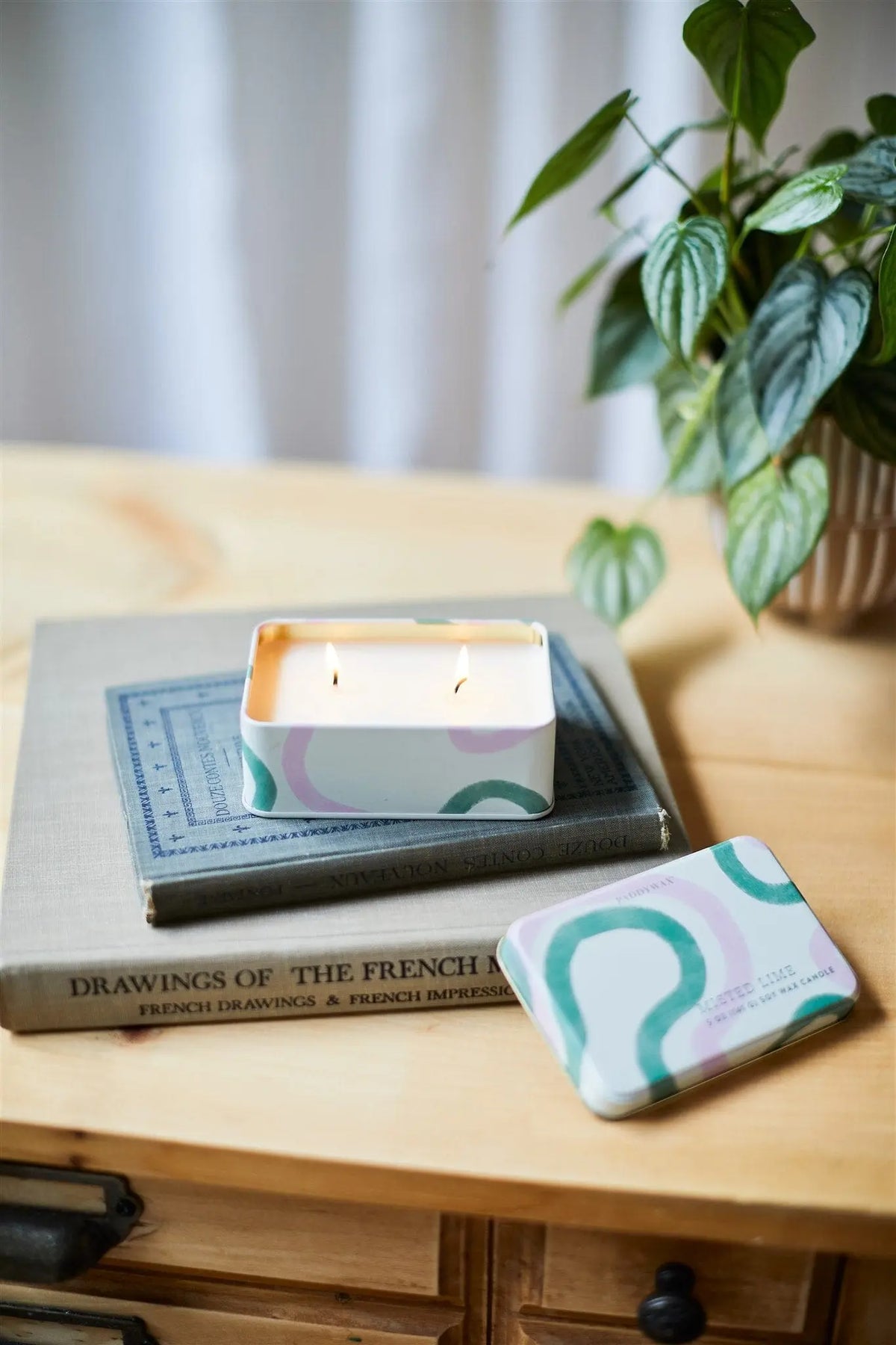 The Misted Lime - 5 oz Tin Candle glows atop stacked books beside its lid, with a potted plant behind on a wooden surface—ideal for freshening up your everyday candle collection.