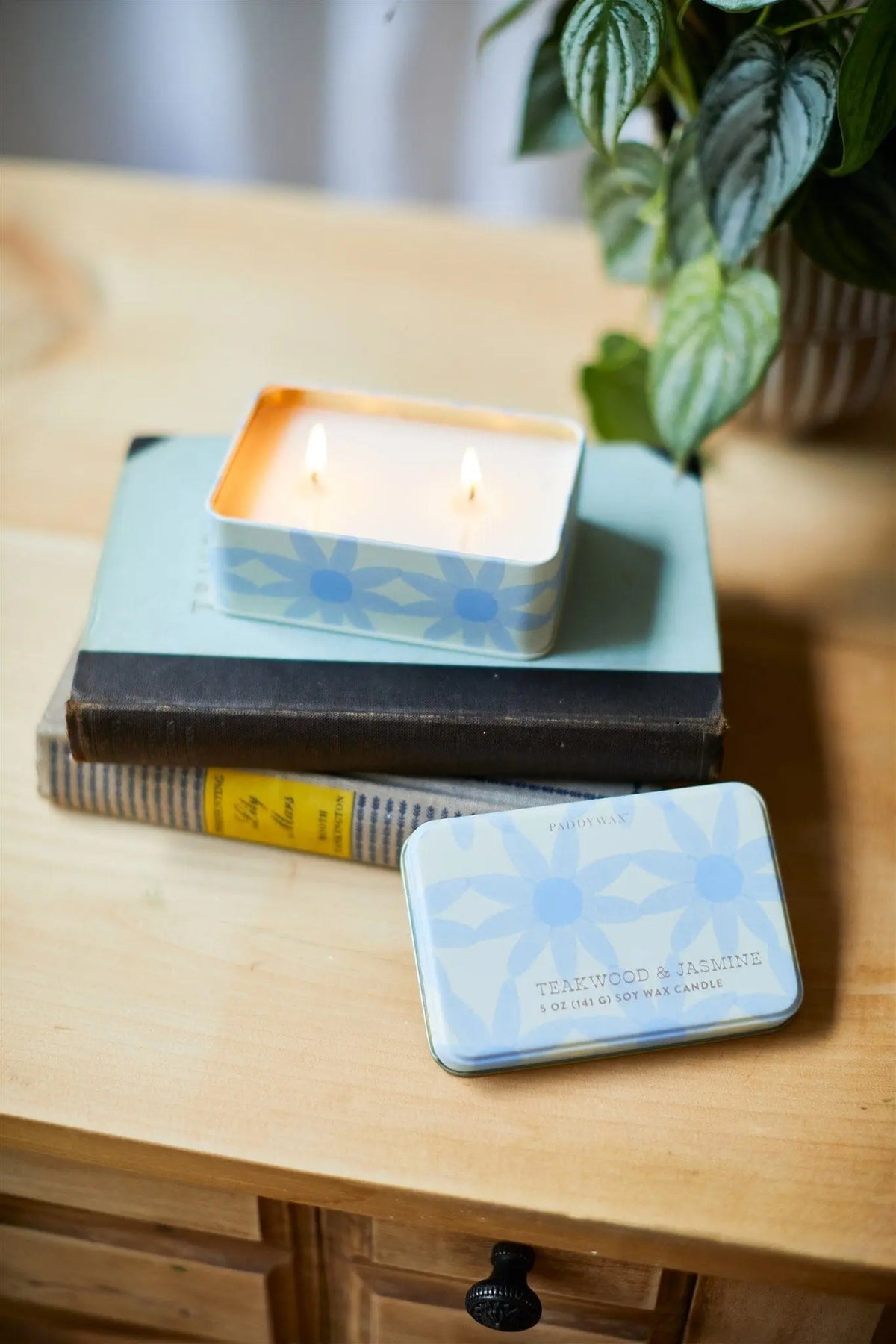 A Teakwood & Jasmine 5 oz Tin Candle with a blue patterned tin sits lit on stacked books next to its lid on a wooden table, with a leafy potted plant nearby.