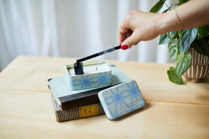 A hand with red nail polish uses the Black Candle Snuffer to extinguish candles in a blue floral tin, set on stacked books beside a potted plant on a wooden surface.