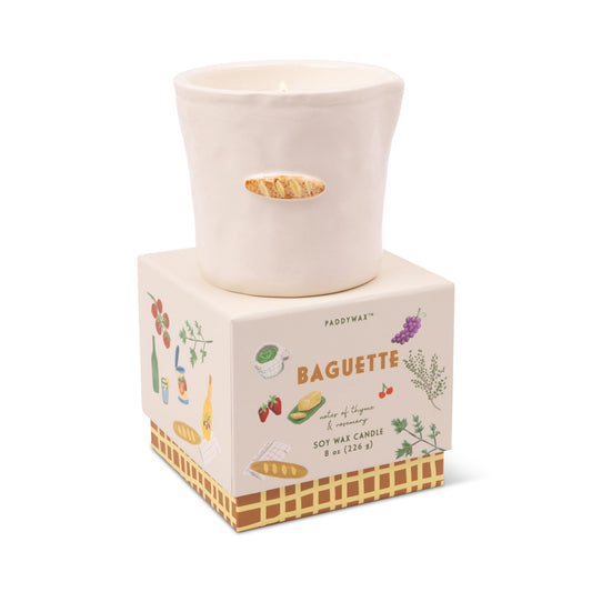 The Baguette - Bistro 8 oz Candle by Paddywax is a white ceramic holder with a small baguette design, presented on a decorative "Baguette" box featuring flowers, bread, and food art. It contains aromatic soy wax.