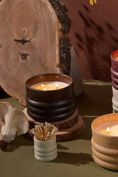 Three lit Tobacco & Patchouli - Grain 12 oz candles in ribbed ceramic holders are displayed on a table with matches, a wooden slab, a mushroom, and a pinecone in the background.