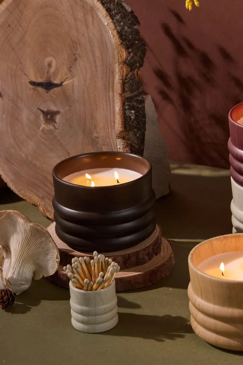 Three lit woodgrain candles in ribbed ceramic holders sit on a green surface near a White Washed Ceramic Wood Match Holder, a wooden log slice, and a mushroom.