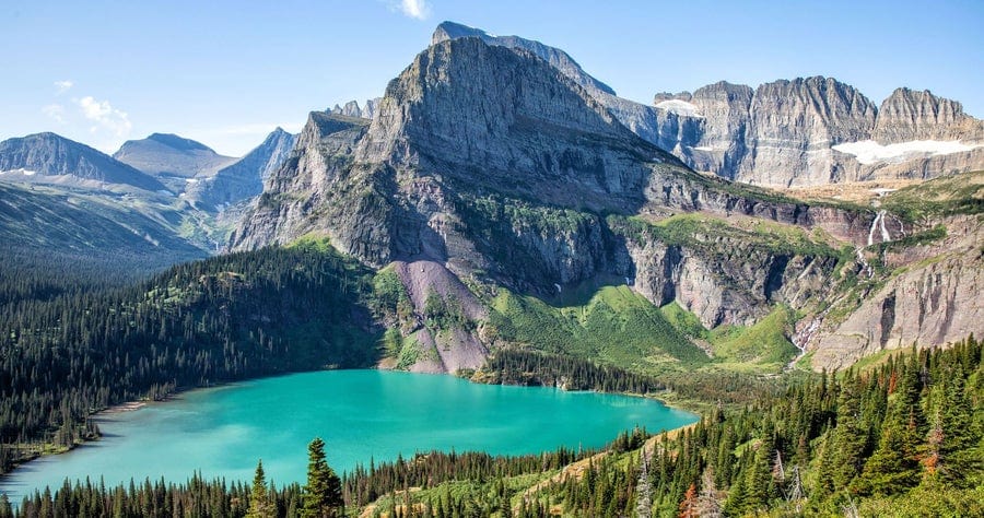 A bright turquoise lake surrounded by dense pine forests sits below tall, rugged mountains with rocky peaks under a clear blue sky.