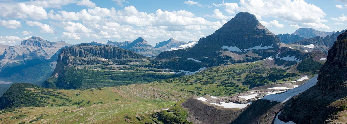 Panoramic view of a mountain range with rugged peaks, patches of snow, green valleys, and scattered clouds in a blue sky. The landscape is expansive and dramatic.