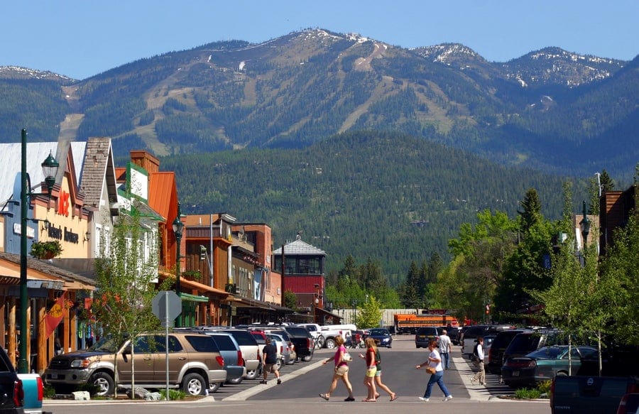 Pedestrians cross a busy street lined with cars and colorful shops, set against a backdrop of green forests and towering mountains under a clear blue sky.