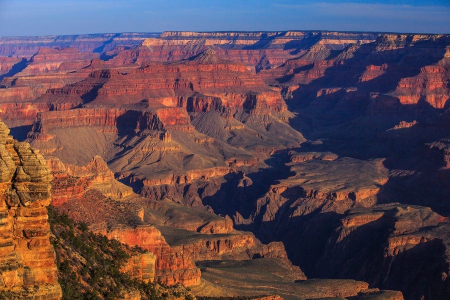A panoramic view of the Grand Canyon at sunset, showcasing steep, layered red and orange rock formations and deep shadows cast across the vast, rugged landscape.