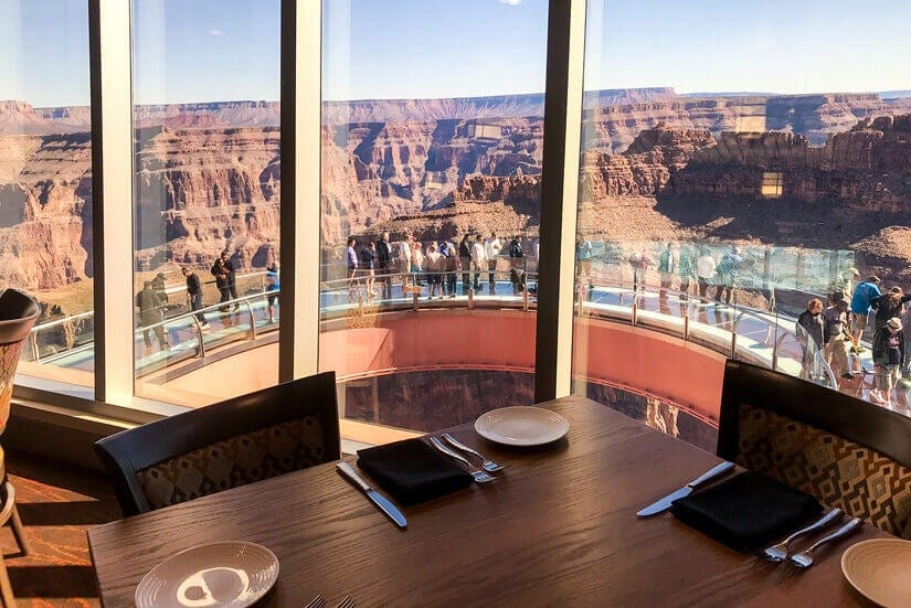 A restaurant table set for four sits by large windows overlooking the Grand Canyon Skywalk, where people are walking along the glass bridge with canyon views in the background.