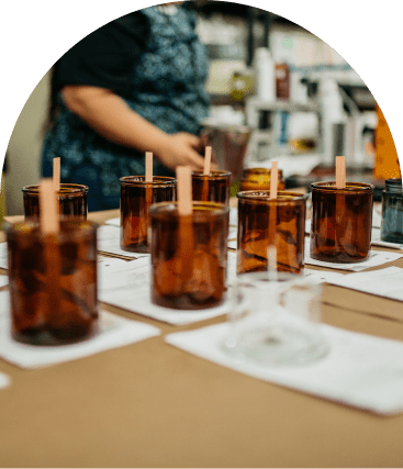 Several amber glass jars with wooden sticks are arranged on a table, likely for candle making, with a person standing in the background.