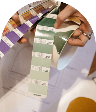 A person holds a ceramic mug and a Pantone color swatch book with various shades of green, above architectural plans on a table.