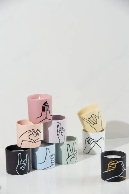 The Lavender + Thyme "Peace" - Impressions 5.75 oz Candle features a vibrant matte holder adorned with a hand gesture illustration, arranged neatly on a white surface.
