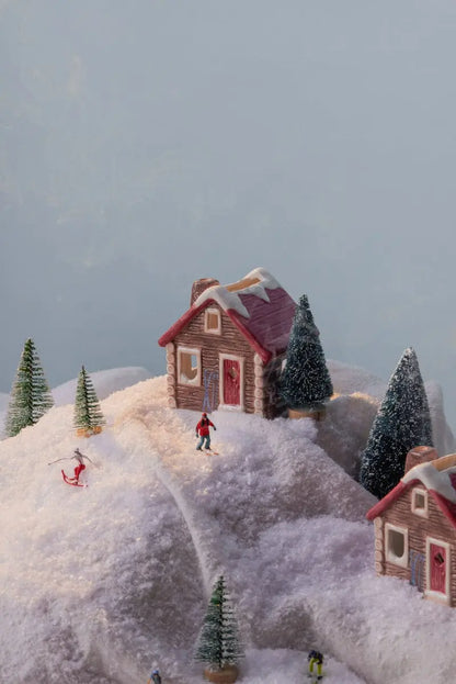 Experience the charm of No. 05 The Alpine Chalet – Incense & Tea Light Holder, featuring a picturesque snowy village scene with petite houses, pine trees, and playful figurines skiing and sledding over pristine white hills set against a soft blue backdrop. This delightful winter display serves as both a ceramic chalet décor piece and a functional tea light or incense holder, infusing any space with warmth and whimsical alpine charm.