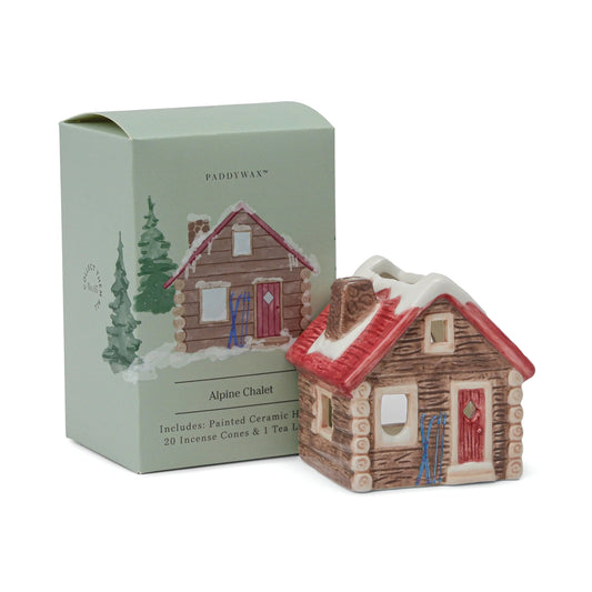 No. 05 The Alpine Chalet - Incense & Tea Light Holder is a charming painted ceramic house, designed in the shape of an alpine chalet, shown beside its box adorned with illustrations of the house and trees. This delightful decor set serves as both a tea light and incense holder, and includes 20 incense cones and a tea light.