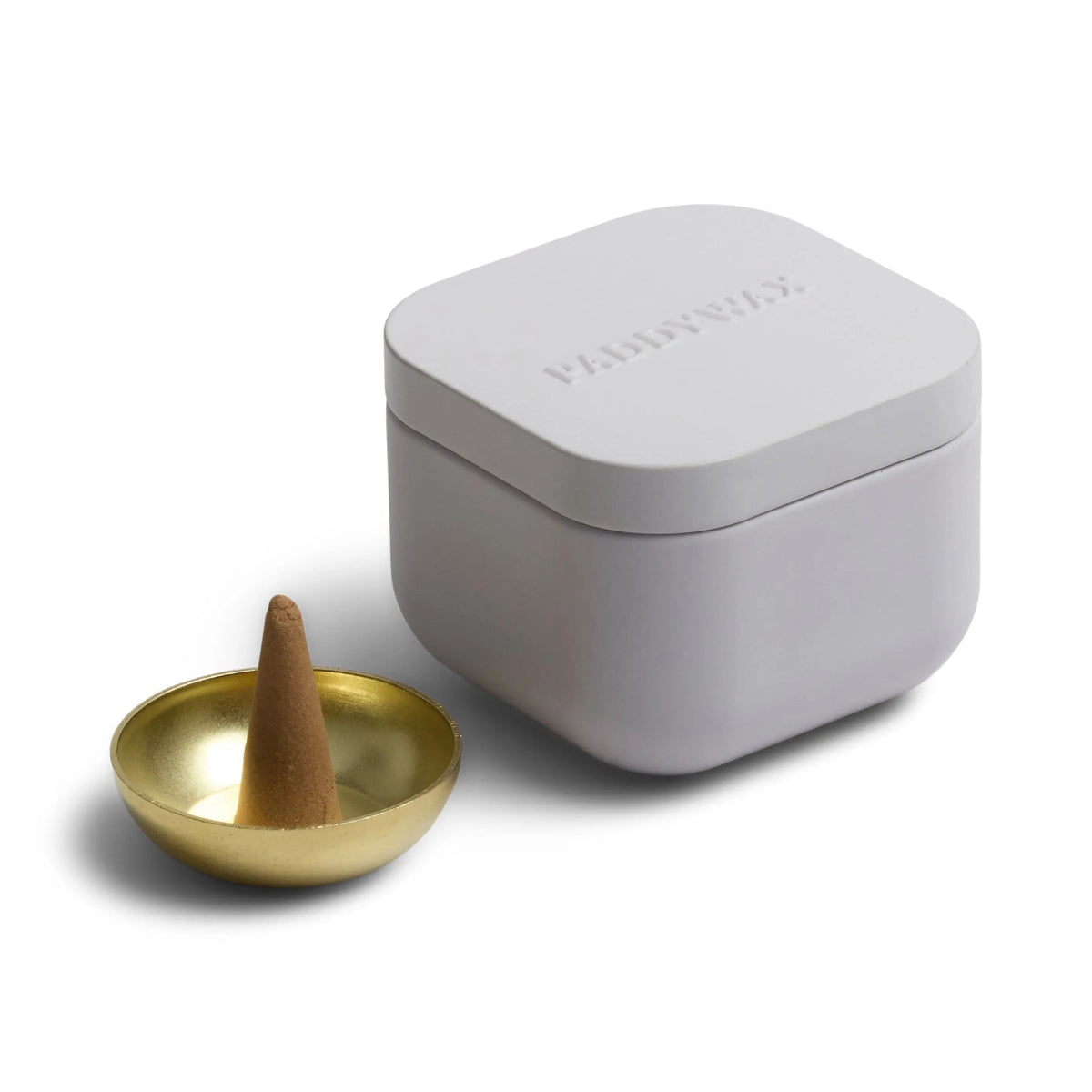 A white, square Cotton & Teak - Metal Incense Tin with a lid sits next to a small gold incense cone holder, which holds a brown incense cone from our collection of top-selling fragrances.