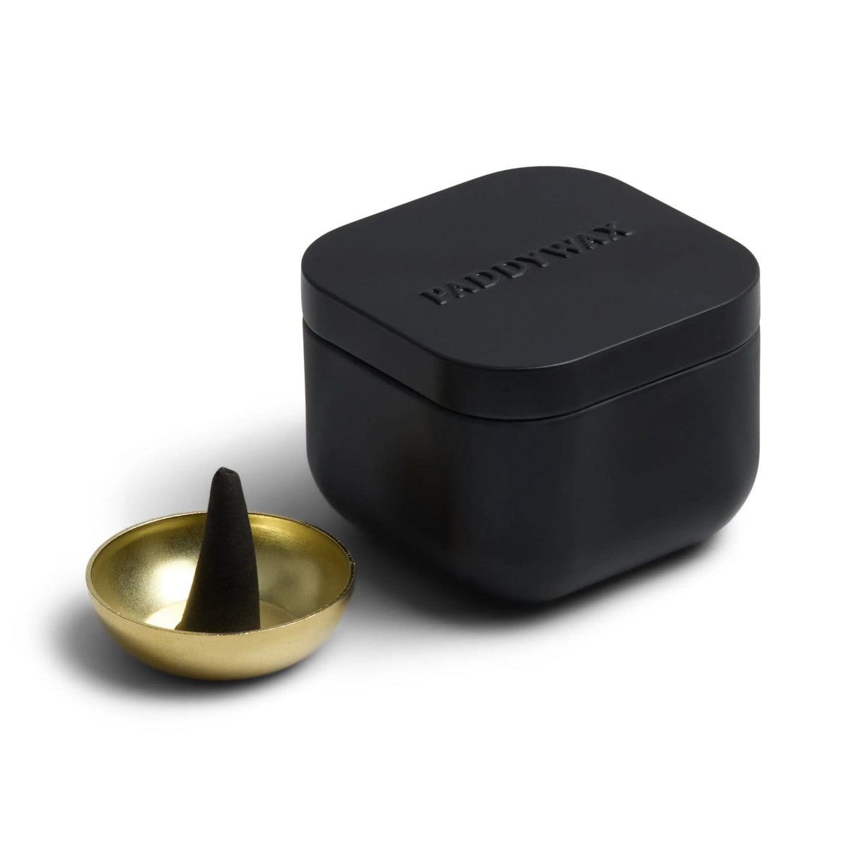 A black box with PADDYWAX embossed on the lid sits beside a small gold incense cone holder, displaying a black Teak & Tobacco - Metal Incense Tin crafted with best-selling fragrances.