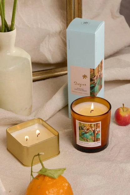 A lit amber glass candle and a lit Degas French Linen + Orris - Impressionist 3.5 oz Tin Candle sit on a beige cloth near a box, vase, orange, and apple. Both candles and the box have art-inspired fragrances with decorative artistic labels.