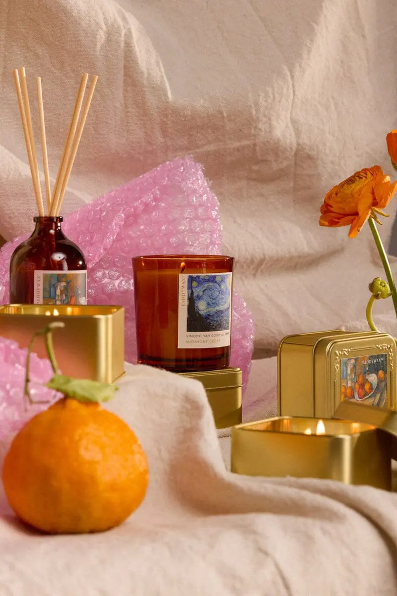 A cozy Vincent Midnight Cedre - Impressionist 6 oz Candle in a gold tin is arranged with a glass reed diffuser, an orange, and an orange flower on beige fabric with pink bubble wrap—perfect for art-inspired gifting.