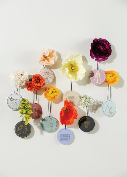 Assorted flowers surround circular embroidered patches with hand gestures and uplifting messages, plus the Mint Leaf + Cardamom "You Got This" Impressions Car Fragrance, all set against a light background.
