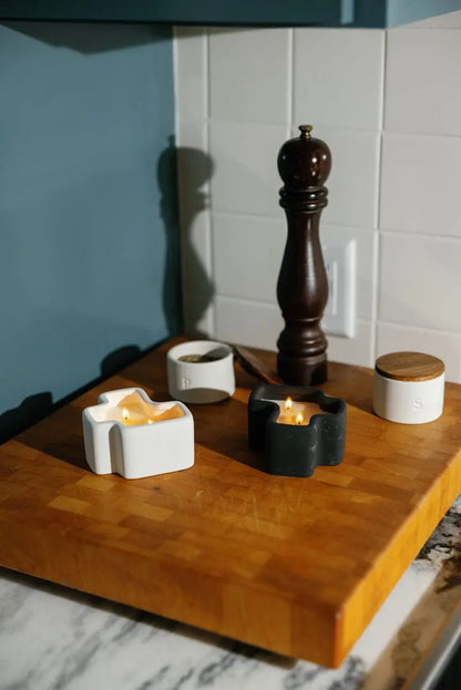A Salted Air & Smoked Woods - Jigsaw 4 oz Candle Boxed Set, featuring two small candles in black and white holders, sits on a wooden cutting board alongside a pepper mill and two small round containers in a kitchen with a white tile backsplash.