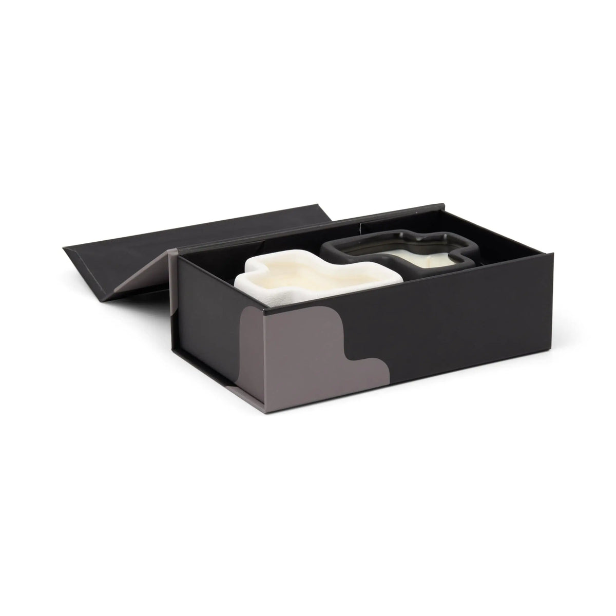 The Salted Air & Smoked Woods - Jigsaw 4 oz Candle Boxed Set features a sleek black box with subtle gray patterns along the sides, designed to complement the jigsaw candle set's minimalist aesthetic. With a partially open lid, the box reveals two white ceramic vessels and a stainless steel cookie cutter nestled inside.