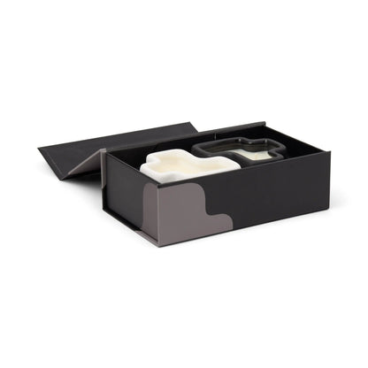 The Salted Air & Smoked Woods - Jigsaw 4 oz Candle Boxed Set features a sleek black box with subtle gray patterns along the sides, designed to complement the jigsaw candle set's minimalist aesthetic. With a partially open lid, the box reveals two white ceramic vessels and a stainless steel cookie cutter nestled inside.