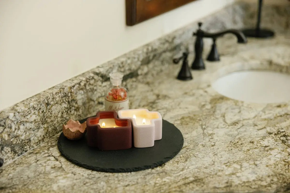 Three lit candles—including the Pink Pistachio & Black Fig Jigsaw 4 oz Candle from a boxed set—a small decorative jar, and a piece of cracked shell rest on a round slate tray atop a marble bathroom counter near a sink with black fixtures.
